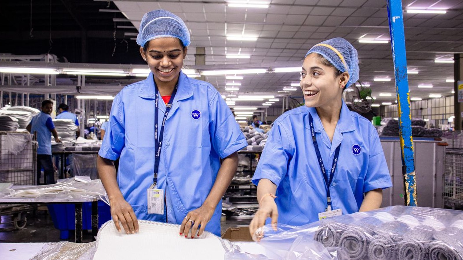 Zwei lachende Mitarbeiterinnen in blauer Arbeitskleidung verpacken Ware in einer Produktionshalle